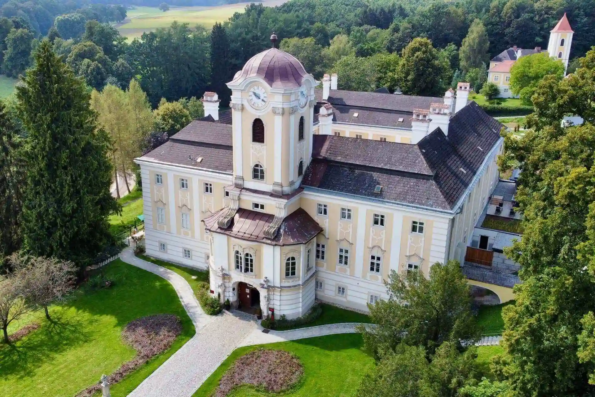 Luftaufnahme eines großen, verschnörkelten, blassgelben Herrenhauses mit einer zentralen Kuppel, umgeben von üppigen grünen Rasenflächen, Bäumen und einem gewundenen Weg in einer ländlichen Landschaft.