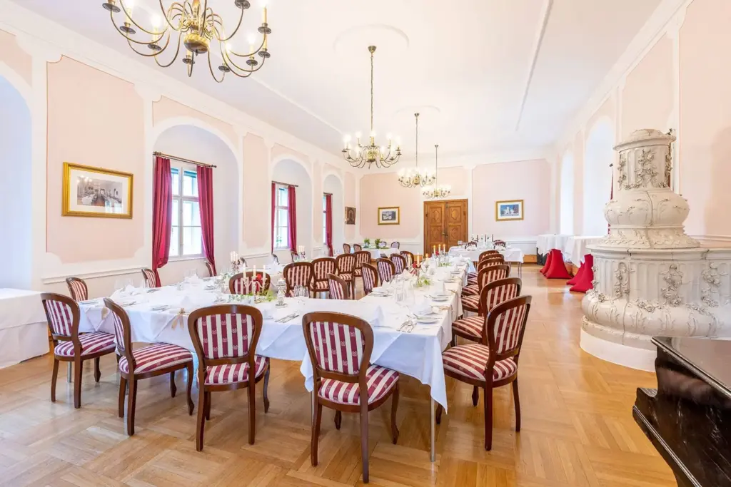 Eleganter Speisesaal mit langen Tischen für ein formelles Essen, umgeben von Holzstühlen mit rot-weiß gestreiften Polstern, Kronleuchtern über dem Kopf, großen Fenstern und zartrosa Wänden mit weißer Zierleiste.