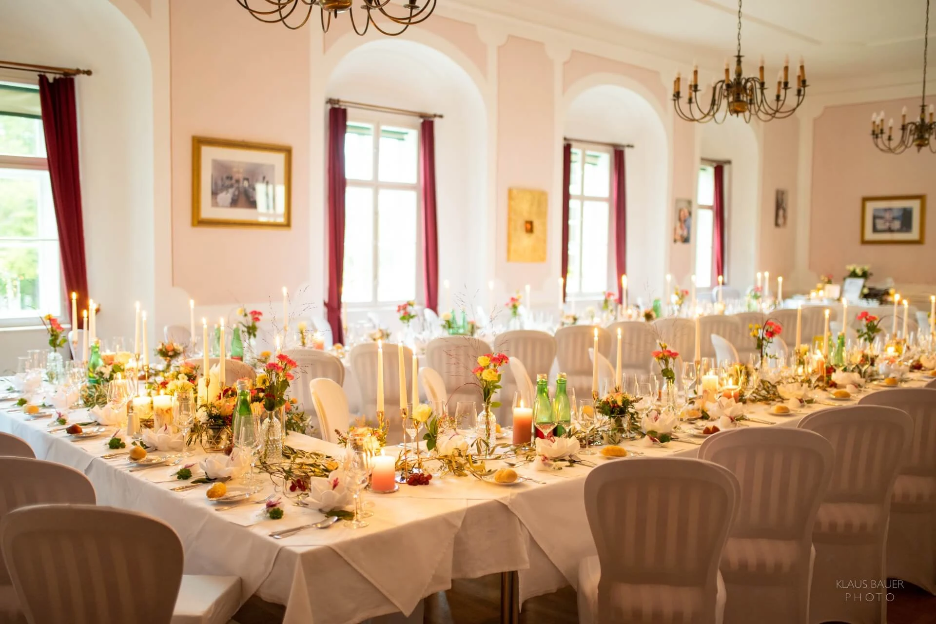 Eleganter Bankettsaal mit weißen Tischtüchern, dekoriert mit Kerzen, Blumen und Tafelgeschirr. Große Fenster lassen natürliches Licht herein; von der Decke hängen Kronleuchter. Die Stühle sind ordentlich um lange, gedeckte Tische herum angeordnet.