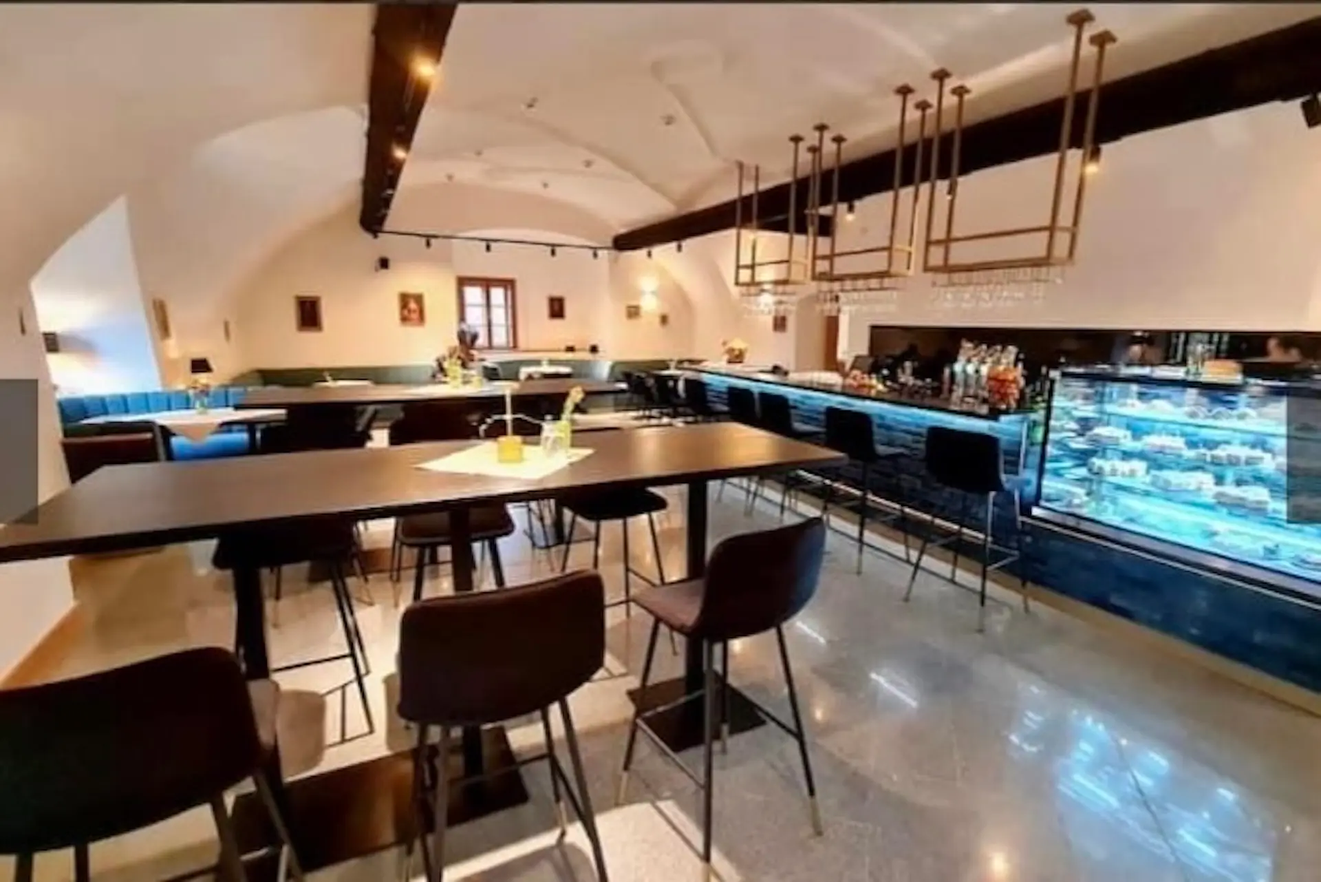 Modernes Café-Interieur mit hohen Tischen und Barhockern, dekorativer Beleuchtung, einem langen Bartresen mit Flaschen und einer Konditorei, blauen Sitzmöbeln an den Wänden und einer hellen, sauberen Atmosphäre.