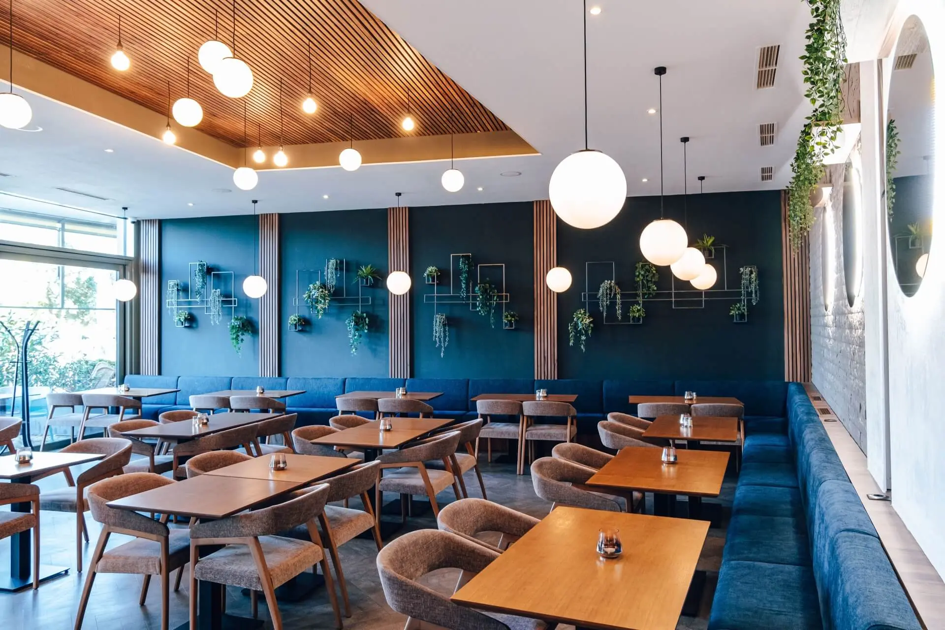 Modernes Restaurantinterieur mit Holztischen und gepolsterten Stühlen, blauer Akzentwand mit geometrischen Regalen und Grünpflanzen, großen Fenstern, hängenden Kugelleuchten und einer Decke mit Holzlatten.