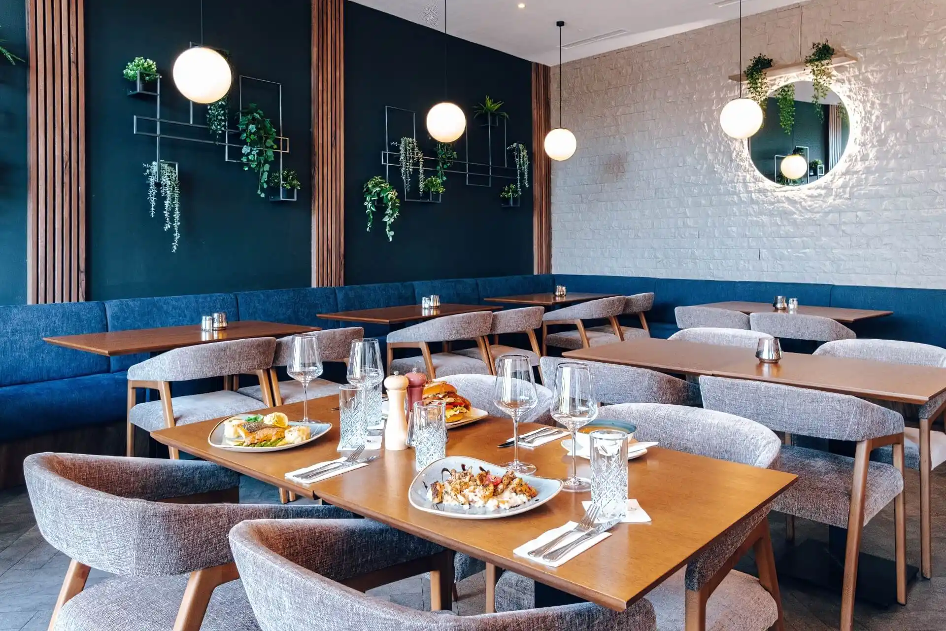 Ein moderner Restaurantbereich mit Holztischen, gepolsterten Stühlen und blauen Sitzbänken. Auf den Tischen stehen Teller mit Speisen und Gläsern. Dekorative Pflanzen und Kugelleuchten hängen an dunklen und hellen Wänden.
