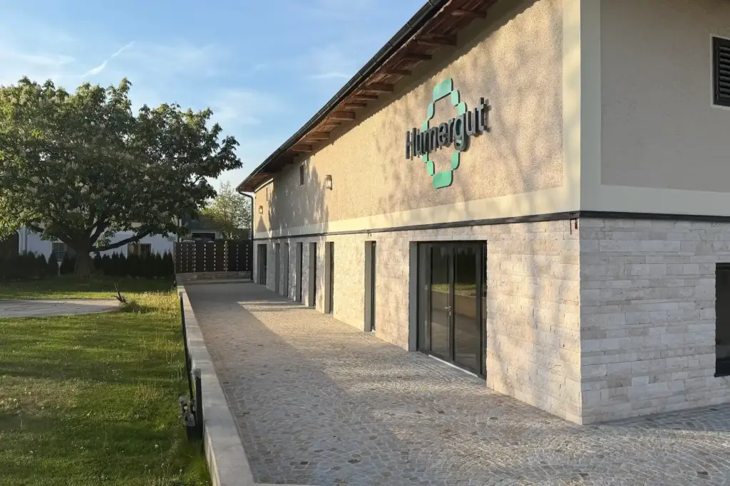 Außenansicht eines Gebäudes mit Steinverkleidung und dem Hinnergut-Logo an der Wand, neben einem mit Steinen gepflasterten Gehweg.