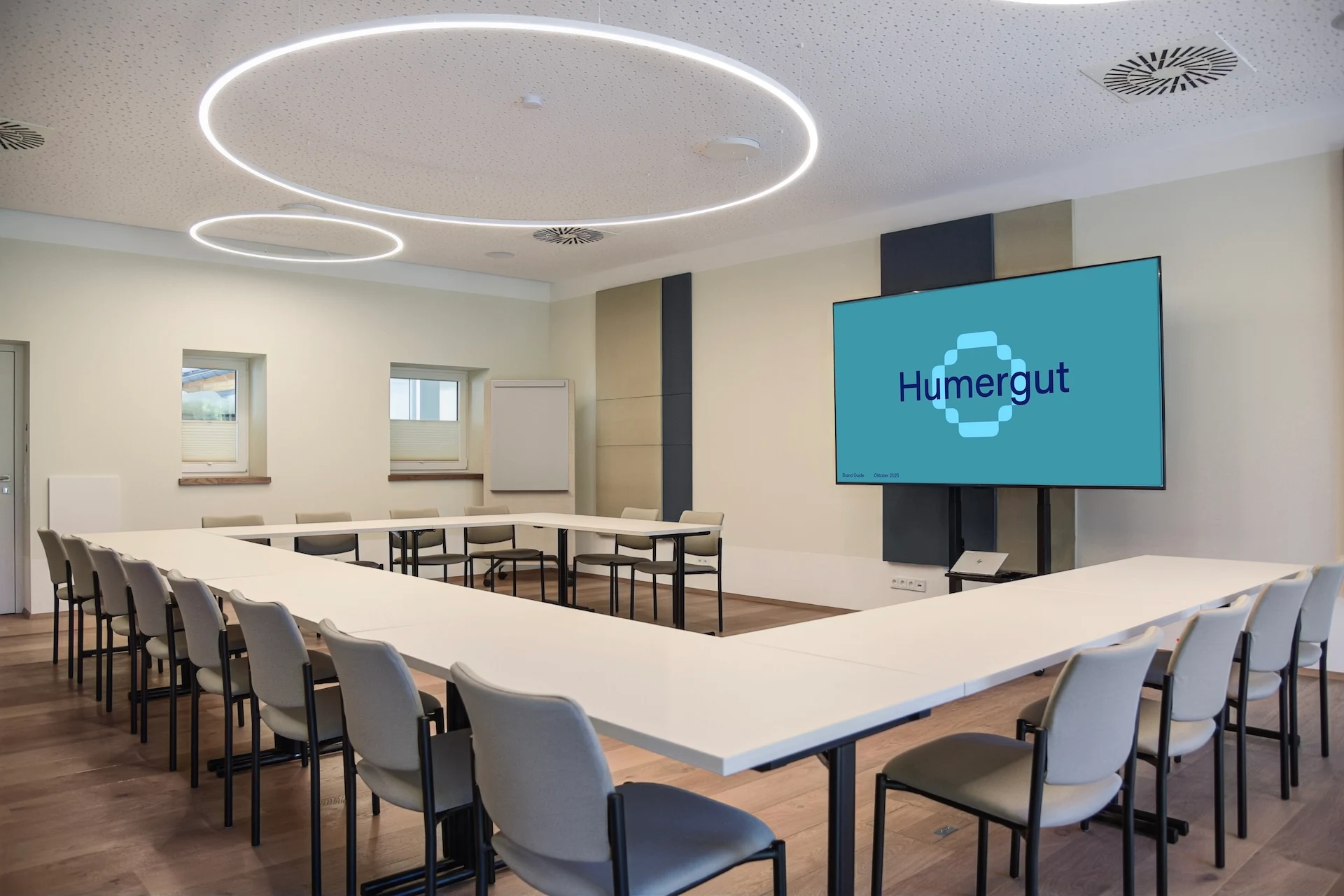 Ein U-förmiger Konferenztisch mit Stühlen in einem Besprechungsraum, gegenüber einer Leinwand, auf der das Humergut-Logo zu sehen ist.