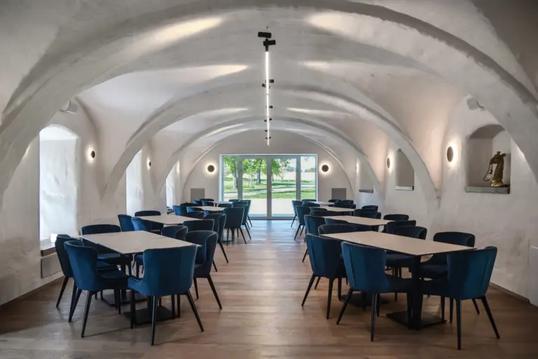 Seminarhof Humergut Essbereich mit Reihen aus blauen Samtstühlen und quadratischen Tischen unter einer gewölbten weißen Decke.
