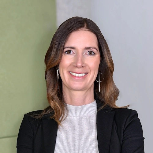 Ein professionelles Porträtfoto einer Frau mit langen braunen Haaren, die einen schwarzen Blazer und ein graues, hochgeschlossenes Oberteil trägt.