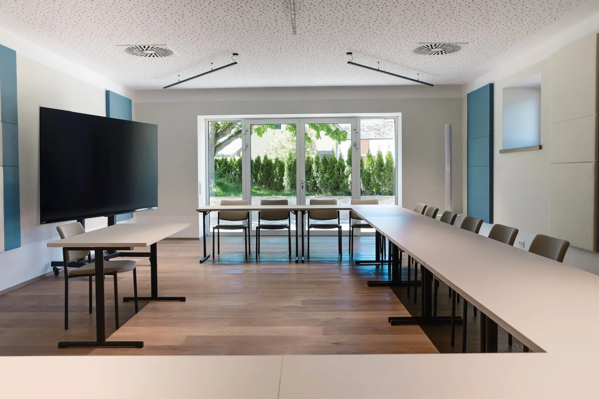 Ein moderner Konferenzraum mit einem großen, an der Wand montierten Monitor und einer U-förmigen Anordnung von Tischen und Stühlen, die auf Glastüren ausgerichtet sind, die zum Garten hin führen.