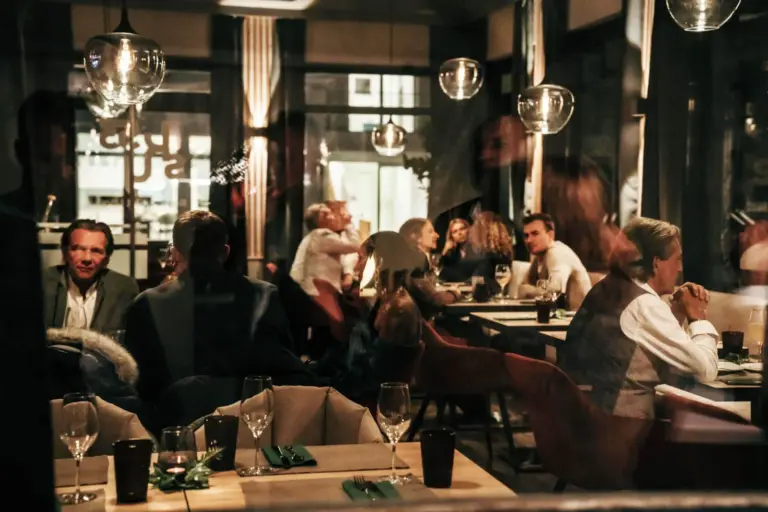 Ein gemütlich beleuchtetes Restaurant, in dem Menschen essen und sich unterhalten. Gläser und Besteck stehen auf den Tischen; die Spiegelungen in den Fenstern sorgen für eine gemütliche, lebendige Atmosphäre.