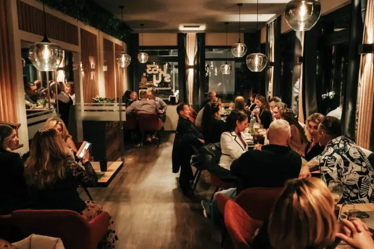 Ein warm beleuchtetes Restaurant mit Gruppen von Menschen, die essen, sich unterhalten und telefonieren. Die moderne Einrichtung, die Hängeleuchten und die großen Fenster sorgen für eine gemütliche, lebendige Atmosphäre am Abend.