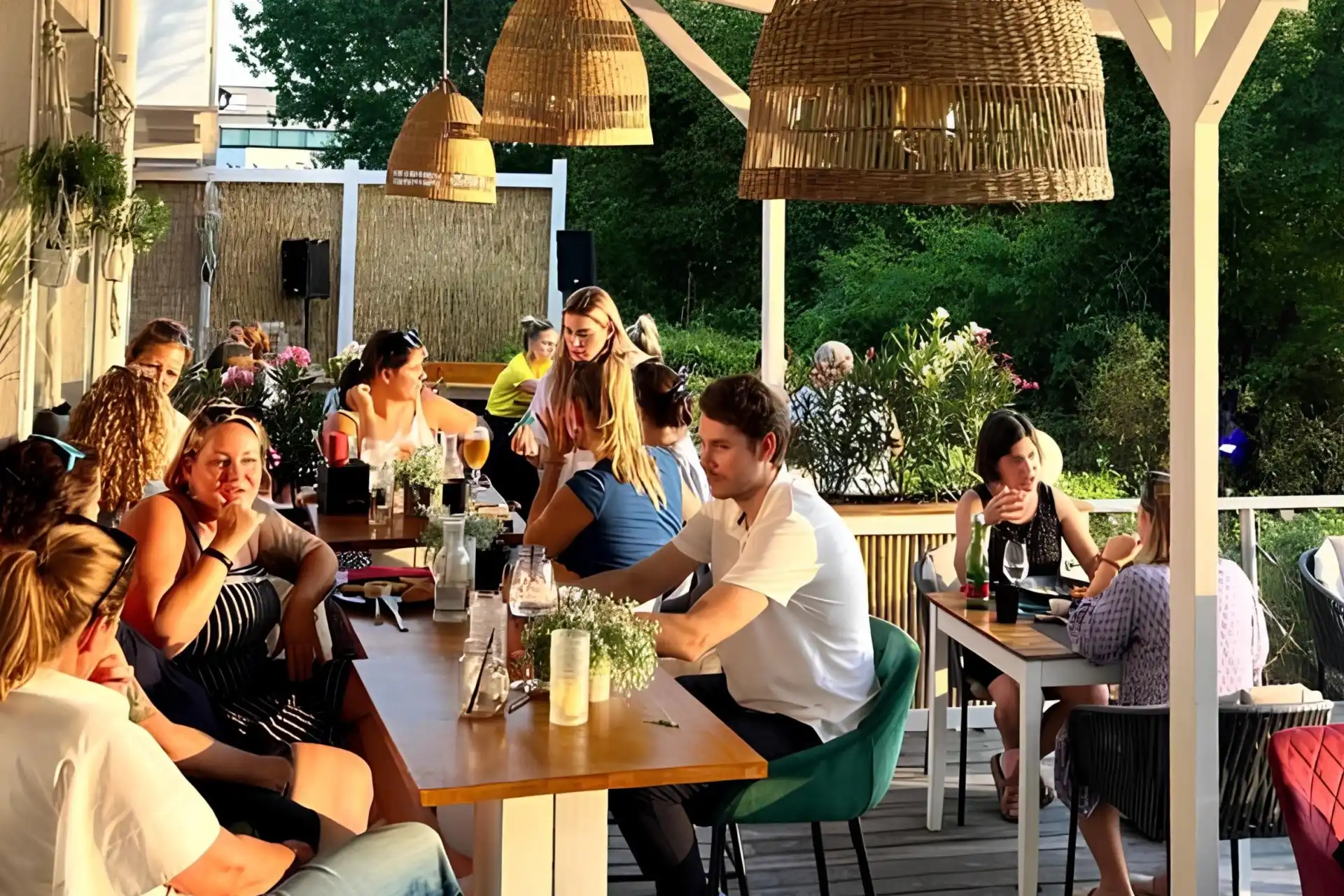 Die Menschen sitzen an Holztischen unter Korblampen auf einer Außenterrasse, plaudern und essen. Üppiges Grün umgibt den Bereich und Sonnenlicht strömt herein und schafft eine warme und lebendige Atmosphäre.