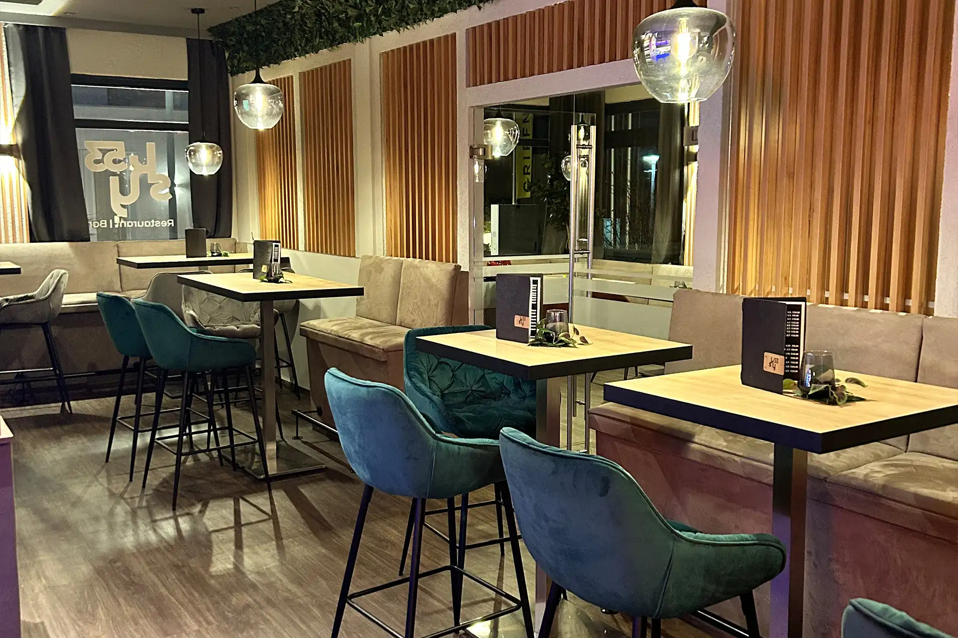Modernes Restaurantinterieur mit Holztischen, tealfarbenen Samtstühlen, beigen gepolsterten Bänken, hölzernen Wandpaneelen, hängenden Glasleuchten und Speisekarten auf jedem Tisch. Der Raum wirkt sauber und stilvoll.