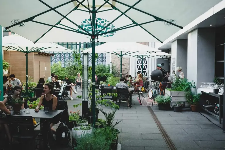 Die Leute sitzen und plaudern an Tischen unter großen Sonnenschirmen auf einer begrünten Terrasse. Die Umgebung ist lebendig, mit Pflanzen und einer entspannten, lockeren Atmosphäre. Einige Leute essen, andere unterhalten sich oder genießen ein Getränk.