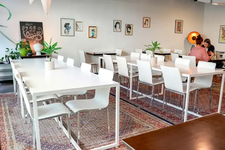 Ein modernes, helles Café mit weißen Tischen und Stühlen, Topfpflanzen, dekorativen Teppichen und gerahmter Kunst an den Wänden. Zwei Leute sitzen in der Ecke und unterhalten sich, während der Rest des Raumes leer ist.