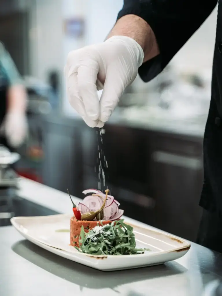 Ein Koch bei Mayer am Pfarrplatz trägt einen weißen Handschuh und streut Salz über ein Gourmetgericht mit frischem Grün und Gemüse, das in einer professionellen Küche elegant auf einem rechteckigen weißen Teller angerichtet wird.