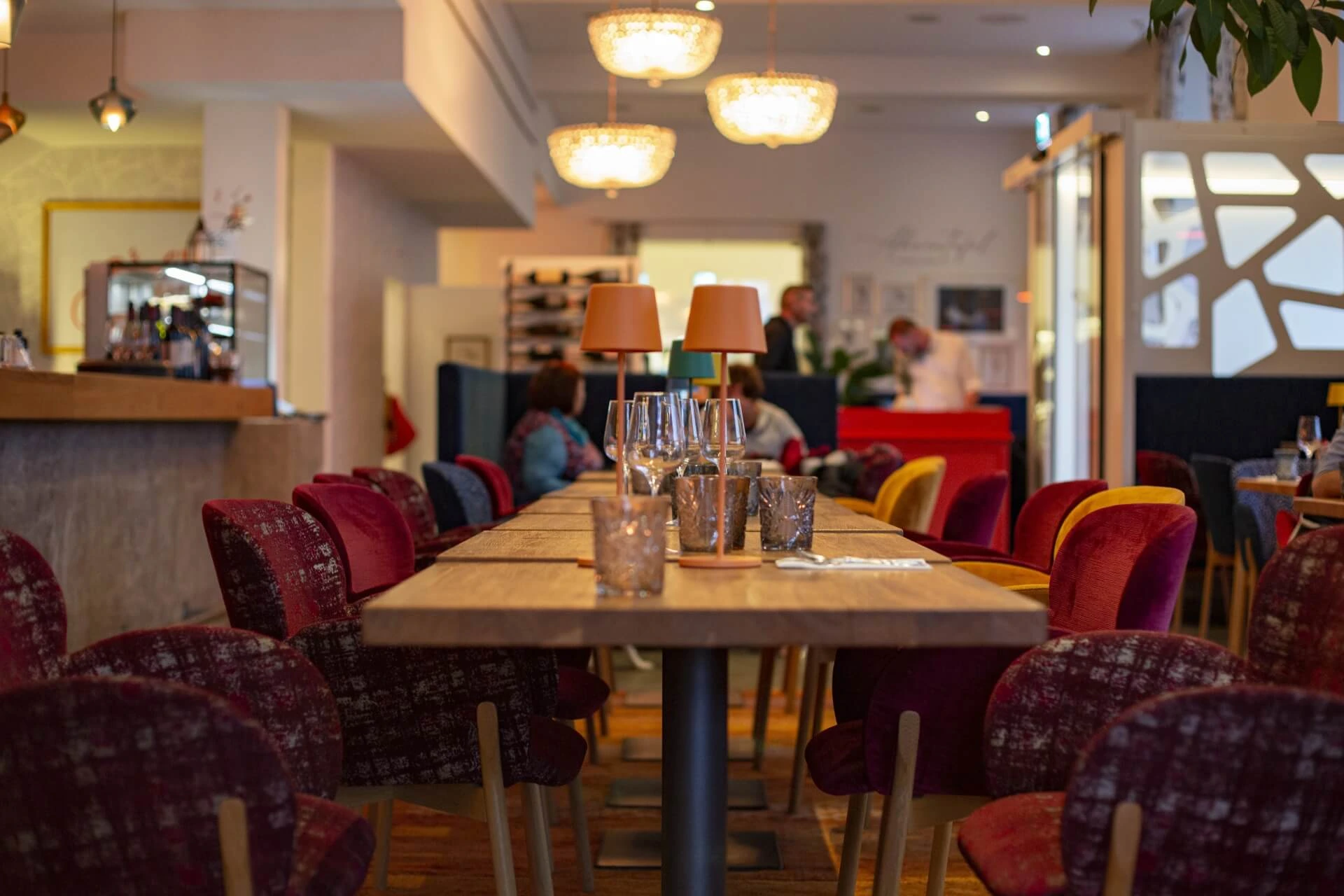 Ein gemütliches Restaurant mit bunten Stühlen, einem langen Holztisch mit Gläsern und Kerzen, warmer Beleuchtung und Menschen im Hintergrund in der Nähe des Eingangs.