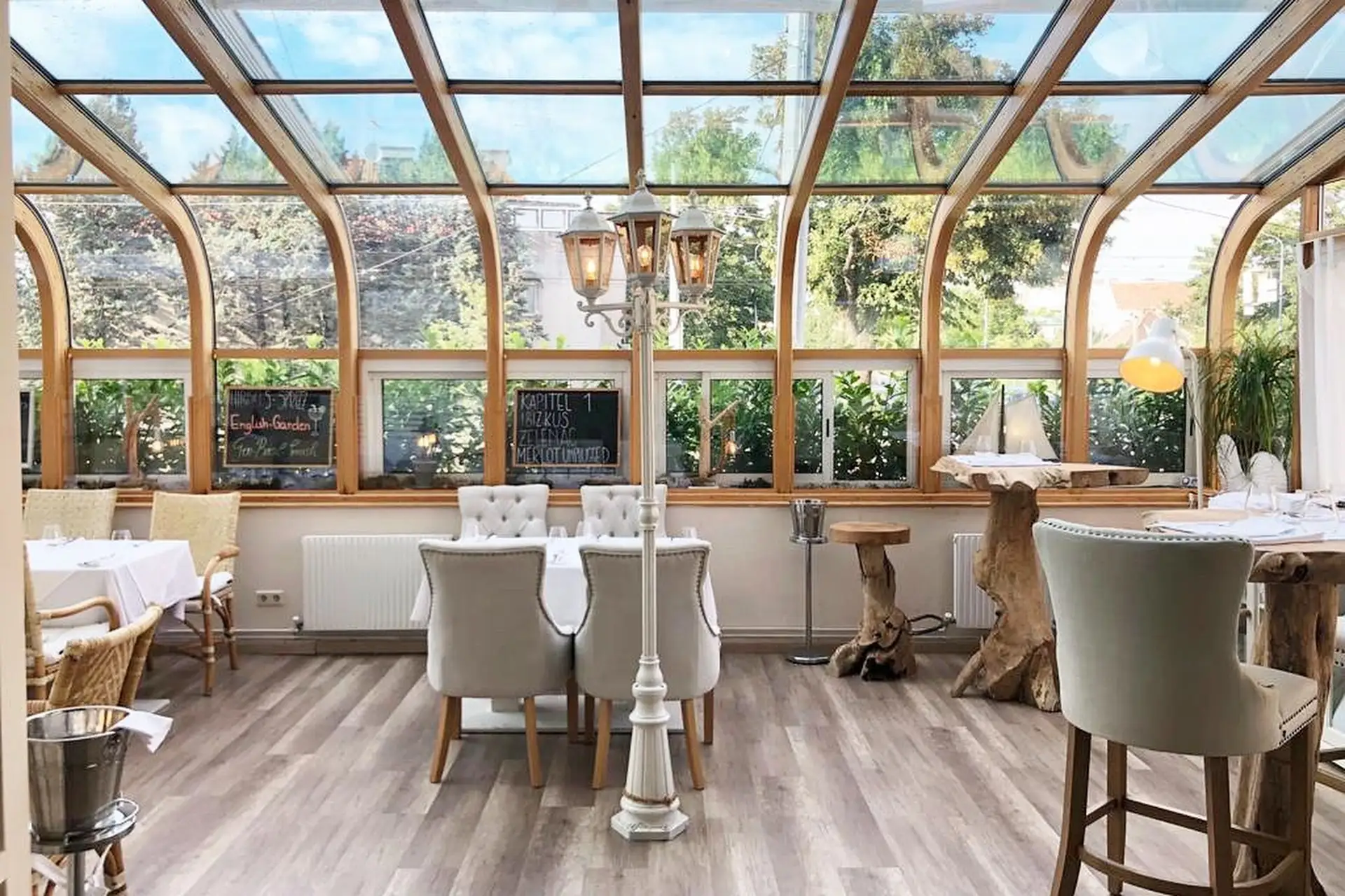 Ein helles, elegantes Restaurant mit Glasdecke und -wänden, weißen Tischdecken, Plüschstühlen, Holzböden und üppigem Grün, das man von außen sehen kann. Der gemütliche Essbereich wird von natürlichem Licht durchflutet.