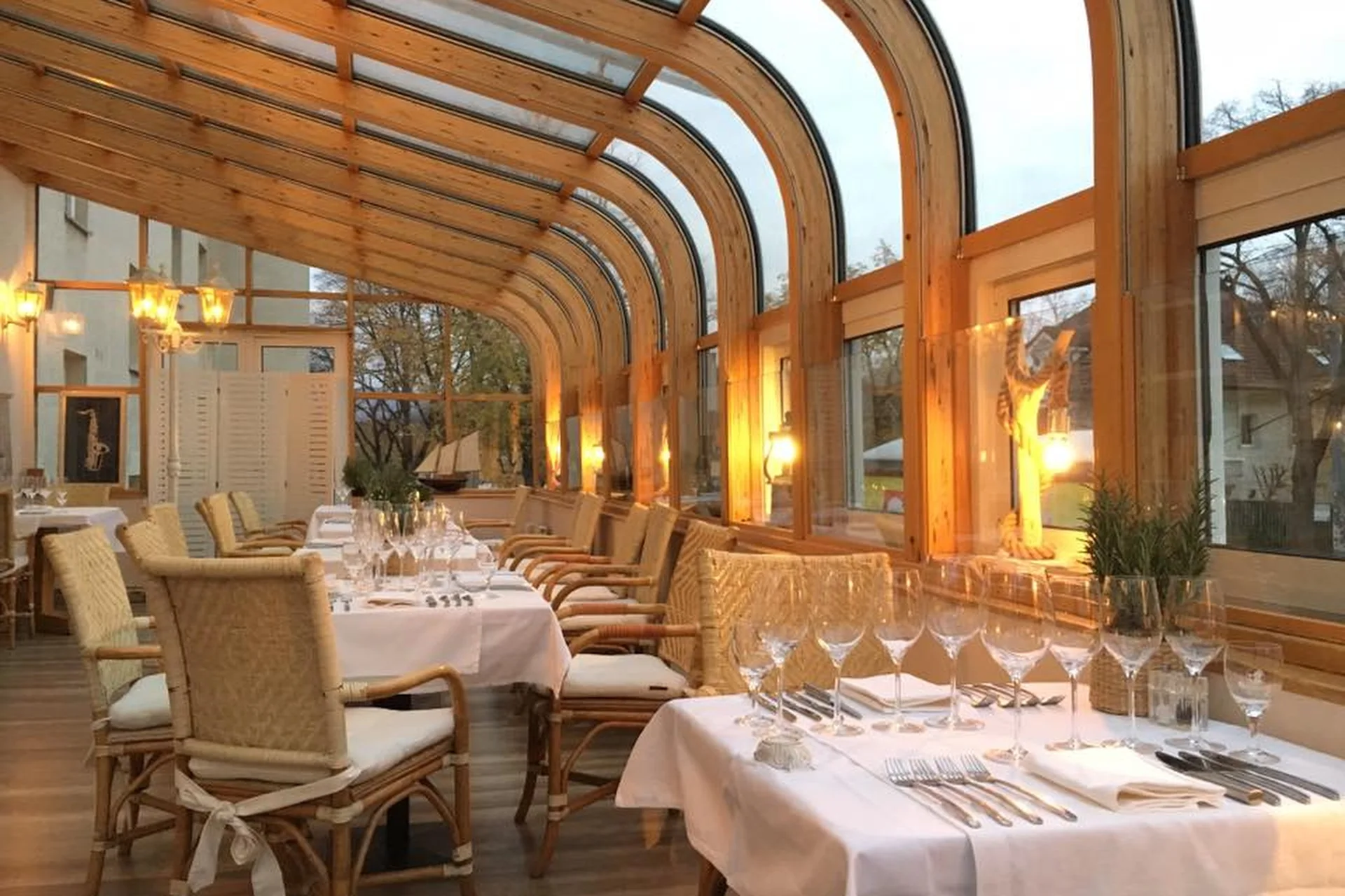 Eleganter Restaurantbereich mit Korbstühlen, weißen Tischtüchern und Weingläsern unter einem geschwungenen Glasdach mit warmer Beleuchtung und großen Fenstern mit Blick auf die Bäume draußen.
