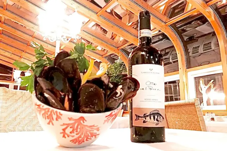 Eine Schale mit gekochten Muscheln, garniert mit Petersilie und Zitrone, steht auf einem Tisch neben einer Flasche Weißwein in einem eleganten, warm beleuchteten Restaurant mit Holzbalken und großen Fenstern im Hintergrund.