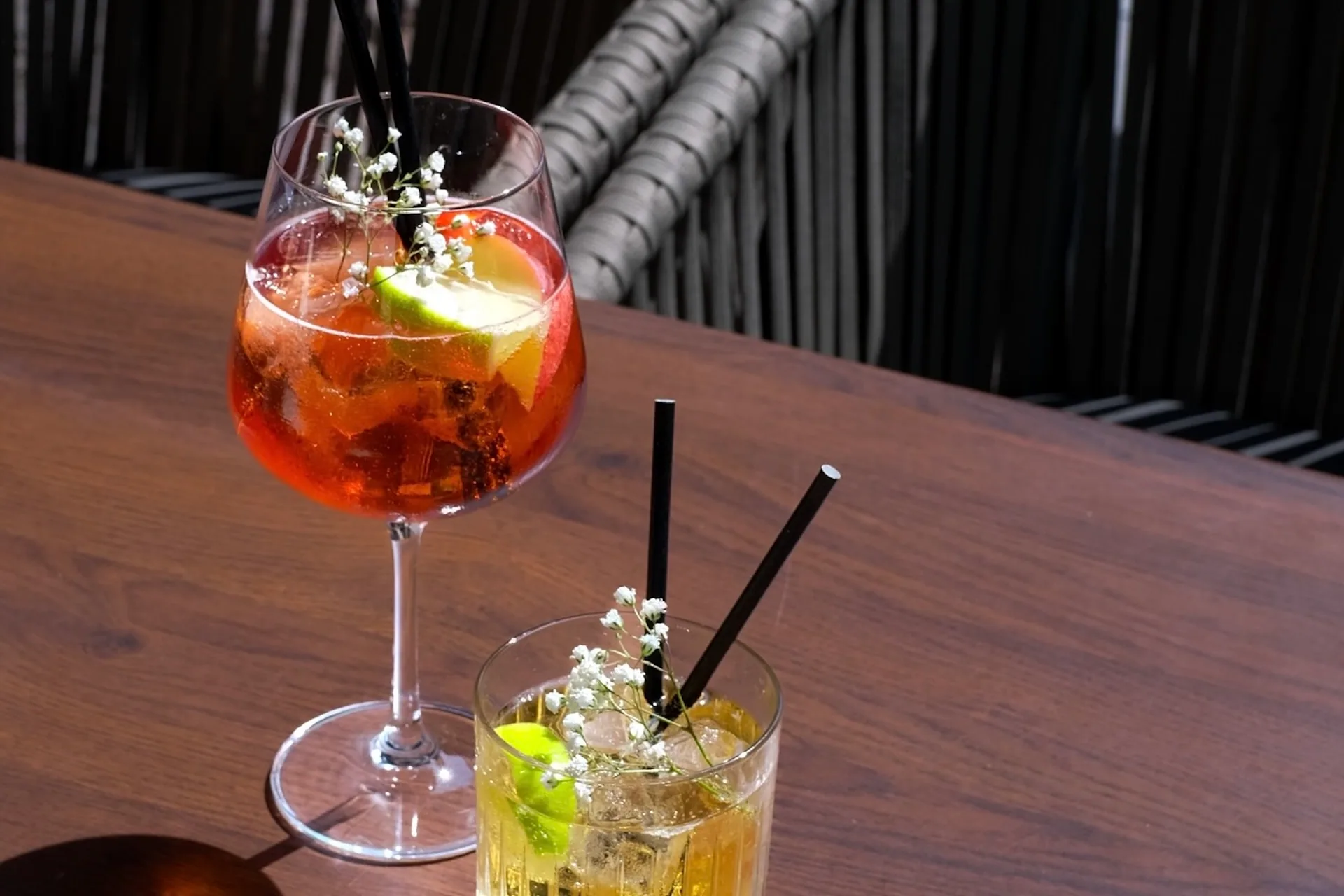 Zwei Cocktails mit Eis, Limettenscheiben und kleinen weißen Blumen stehen auf einem Holztisch; der eine ist rot in einem Weinglas, der andere gelb in einem kurzen Glas. In beiden Getränken befinden sich schwarze Strohhalme.