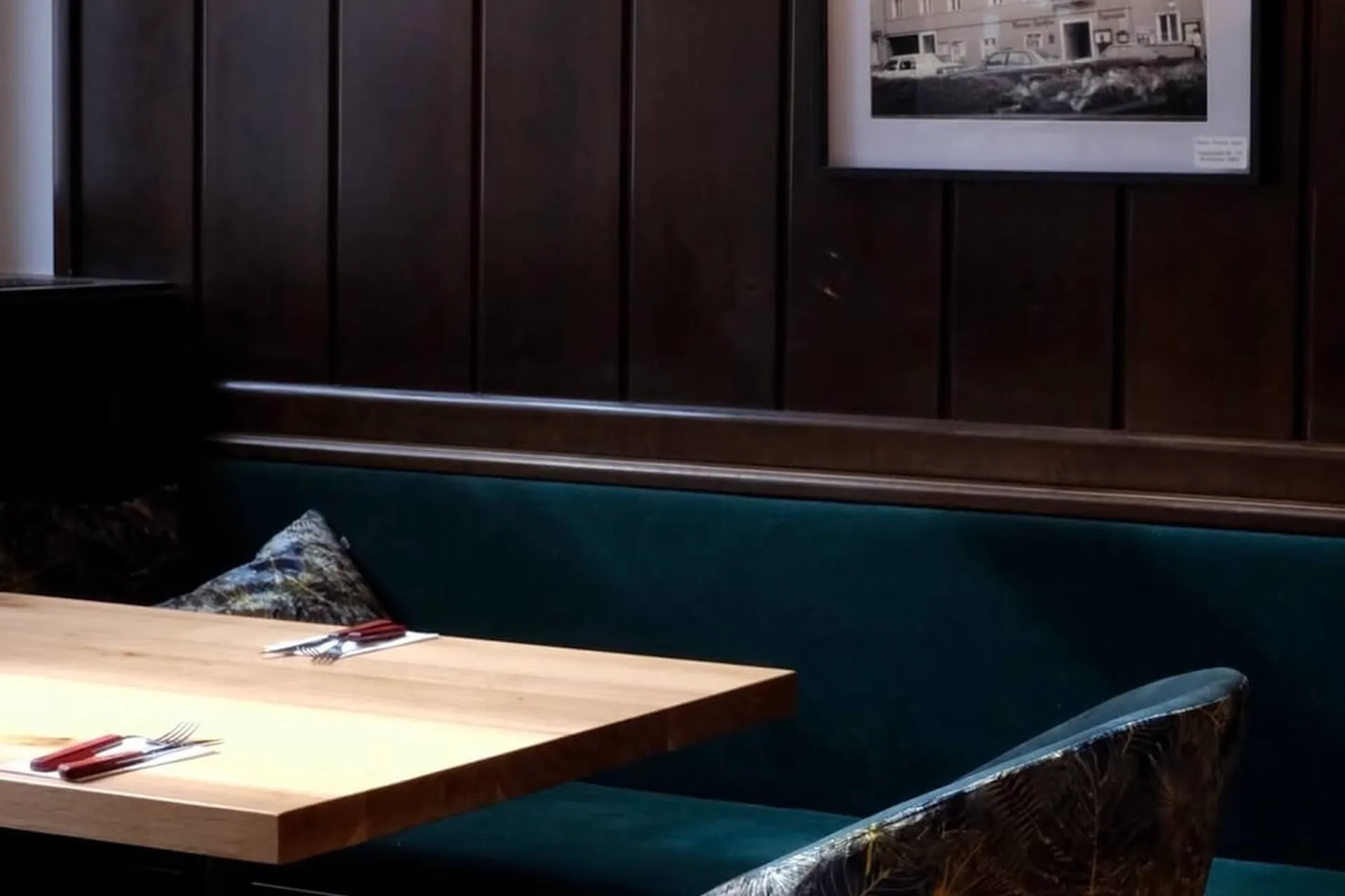 Ein gemütlicher Restaurantstand mit einem Holztisch, dunkelgrünen Sitzpolstern, dekorativen Kissen und ordentlich aufgestellten Utensilien vor einer dunklen holzgetäfelten Wand, über der ein Schwarz-Weiß-Foto hängt.