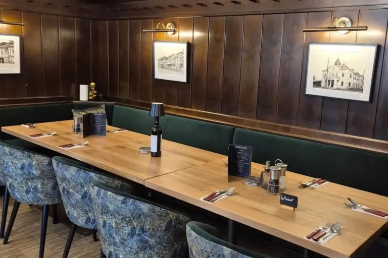 Ein gemütlicher Restaurant-Essbereich mit holzgetäfelten Wänden, gerahmten Schwarz-Weiß-Fotos, einem langen Holztisch für sechs Personen, Speisekarten, Besteck und einer Weinflasche, mit gemusterten blauen Stühlen und grün gepolsterten Sitzbänken.