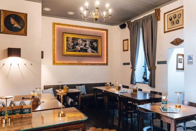 Gemütliches Café mit Holztischen und -stühlen, sanfter Beleuchtung, gerahmter Kunst an weißen Wänden, einem großen Gemälde in der Mitte und Vorhängen an den Fenstern. Die Atmosphäre ist warm und einladend, und auf den Tischen stehen Speisekarten.