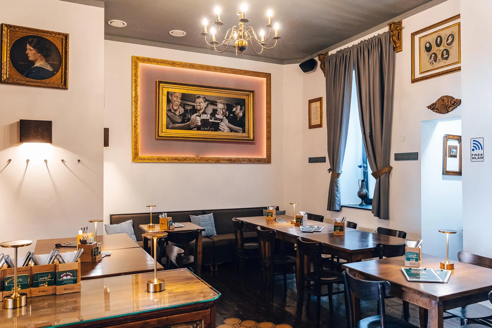 Gemütliches Café mit Holztischen und -stühlen, sanfter Beleuchtung, gerahmter Kunst an weißen Wänden, einem großen Gemälde in der Mitte und Vorhängen an den Fenstern. Die Atmosphäre ist warm und einladend, und auf den Tischen stehen Speisekarten.