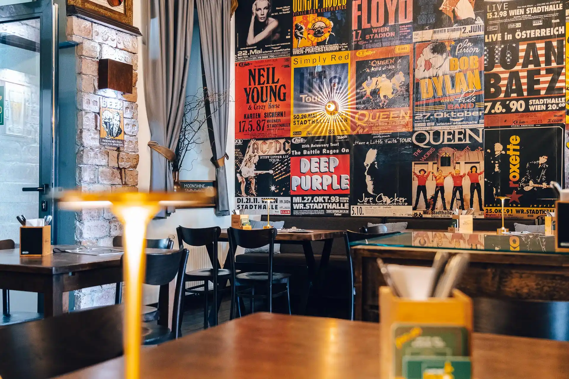 Ein gemütliches Café mit Holztischen und -stühlen und einer bunten Wand mit alten Konzertplakaten von Bands wie Pink Floyd, Queen und Deep Purple. Die warme Beleuchtung schafft eine entspannte Atmosphäre.
