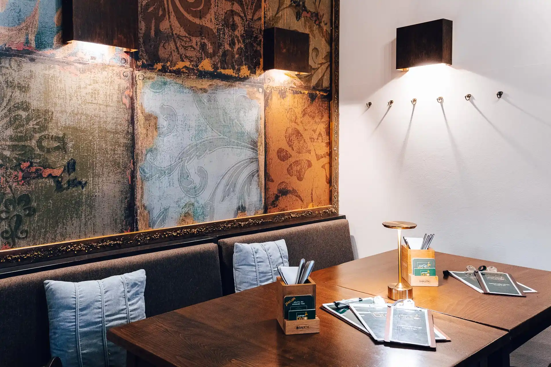 Eine gemütliche Restaurantecke mit einem Holztisch für vier Personen, gepolsterten Sitzbänken, Speisekarten, Besteckhaltern und warmen Wandlampen, die gemusterte Tapeten beleuchten.