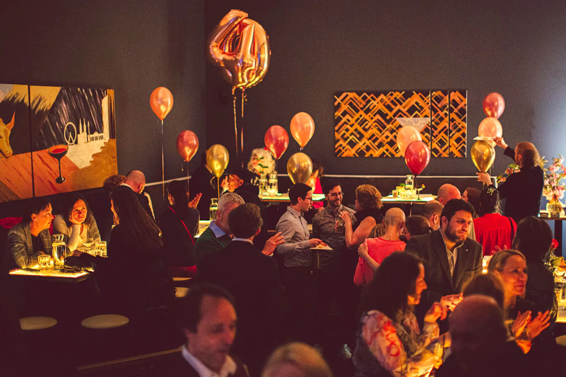 Eine lebhafte Gruppe von Menschen, die sich auf einer schwach beleuchteten Party unterhalten, die mit goldenen und rosafarbenen Luftballons geschmückt ist, darunter ein großer 40er Ballon. An den dunklen Wänden hängt Kunst und die Gäste unterhalten sich an Tischen mit leuchtenden Lichtern.