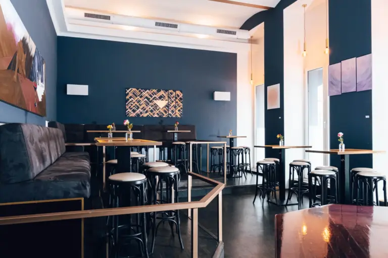 Moderne Café-Einrichtung mit dunkelblauen Wänden, Holztischen und schwarzen Hockern, kleinen Vasen mit Blumen auf den Tischen und abstrakter Kunst an den Wänden. Der Raum ist mit natürlichem Licht und Hängeleuchten gut beleuchtet.