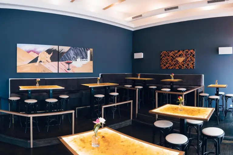 Ein modernes Café-Interieur mit dunklen Wänden, Holztischen, schwarzen Hockern, Sitzbänken und minimalistischer Wandkunst. Kleine Vasen mit einzelnen Blumen schmücken die Tische. Der Raum ist gut beleuchtet und aufgeräumt.