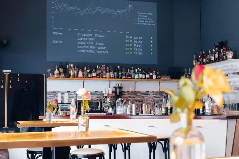Eine moderne Bar mit Flaschen und Gläsern in Regalen, einer Getränkepreisanzeige an der Wand und Tischen mit Blumenvasen im Vordergrund. Die Atmosphäre ist hell und stilvoll.