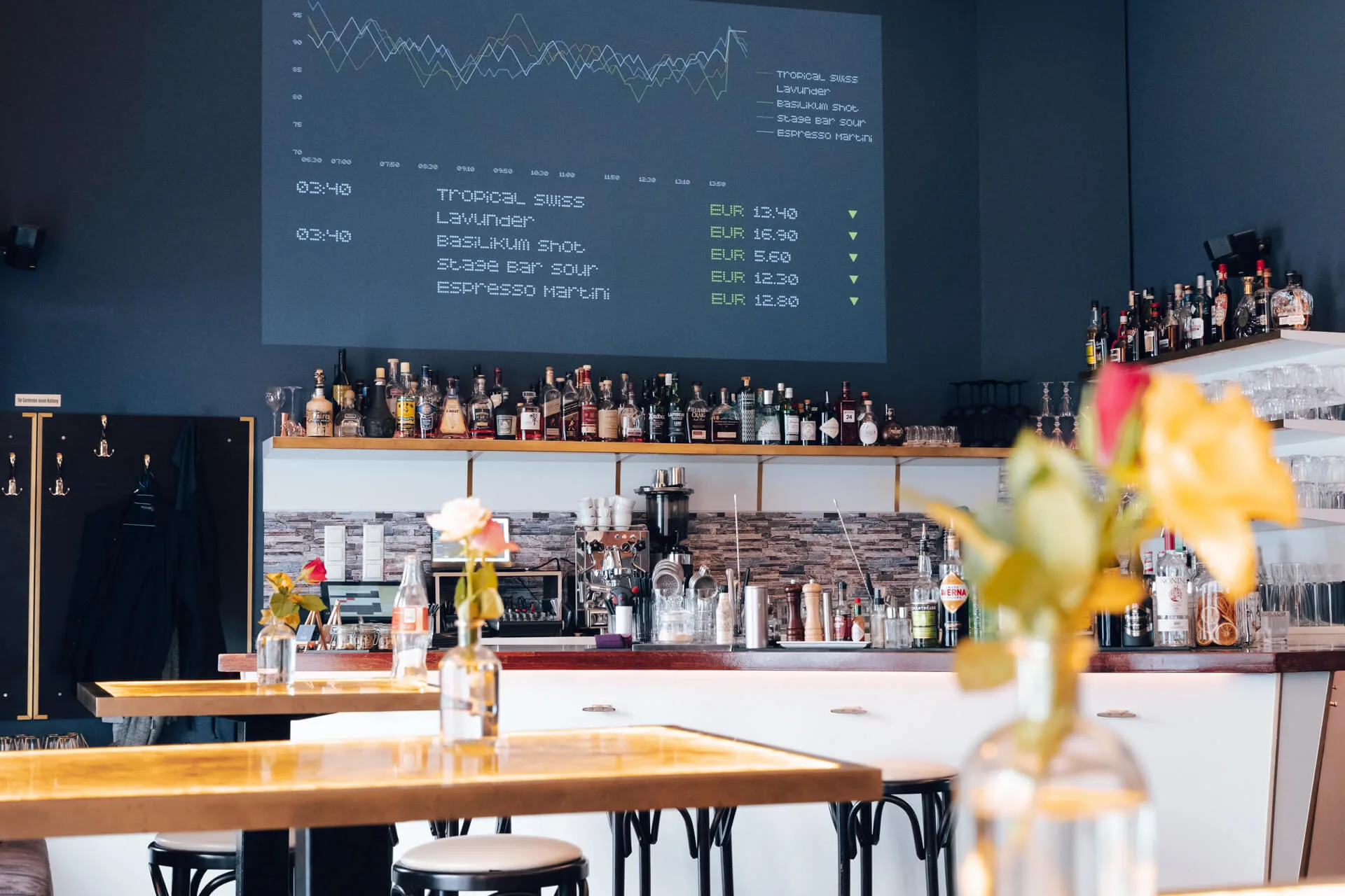 Eine moderne Bar mit Flaschen in Regalen hinter dem Tresen, frischen Blumen auf den Tischen und einer digitalen Speisekarte mit Cocktailpreisen und einem Diagramm an der Wand. Das Ambiente wirkt sauber und stilvoll mit einer entspannten Atmosphäre.