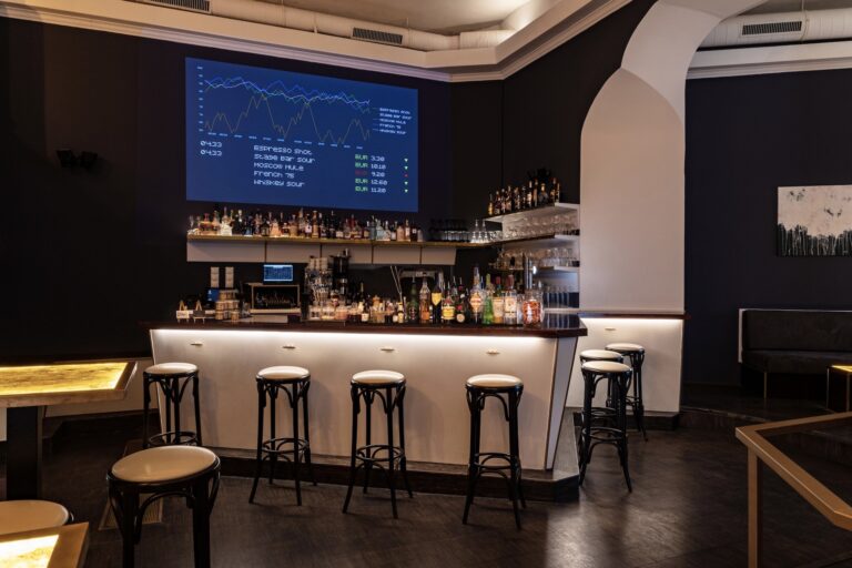 Eine moderne Stage Bar mit einem gut bestückten Tresen, mehreren Barhockern und einem großen digitalen Bildschirm, auf dem bunte Finanzdiagramme und Daten angezeigt werden. Die Bar verfügt über eine gedämpfte Beleuchtung und ein schlichtes, modernes Design.