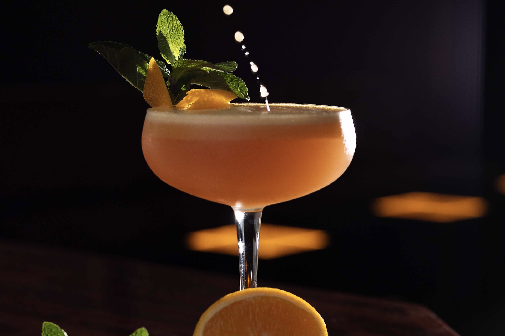 Ein Cocktail in einem Coupe-Glas mit Schaumkrone, garniert mit einem Minzzweig und einer Orangenspirale, fängt die Tropfen auf, die hineinplätschern. Eine Orangenscheibe und Minzblätter liegen daneben und fangen den lebendigen Geist der Stage Bar auf einer dunklen Oberfläche ein.