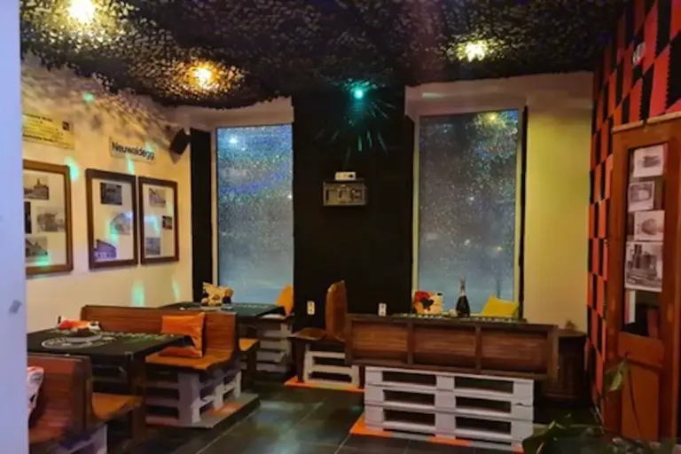 Ein gemütliches Café mit Möbeln aus Paletten, Holzbänken und Tischen. An den Wänden hängen gerahmte Fotos und Kunstwerke, und zwei große Fenster zeigen Regentropfen von draußen. An der Decke befinden sich dunkle Netze und warme, leuchtende Lichter.