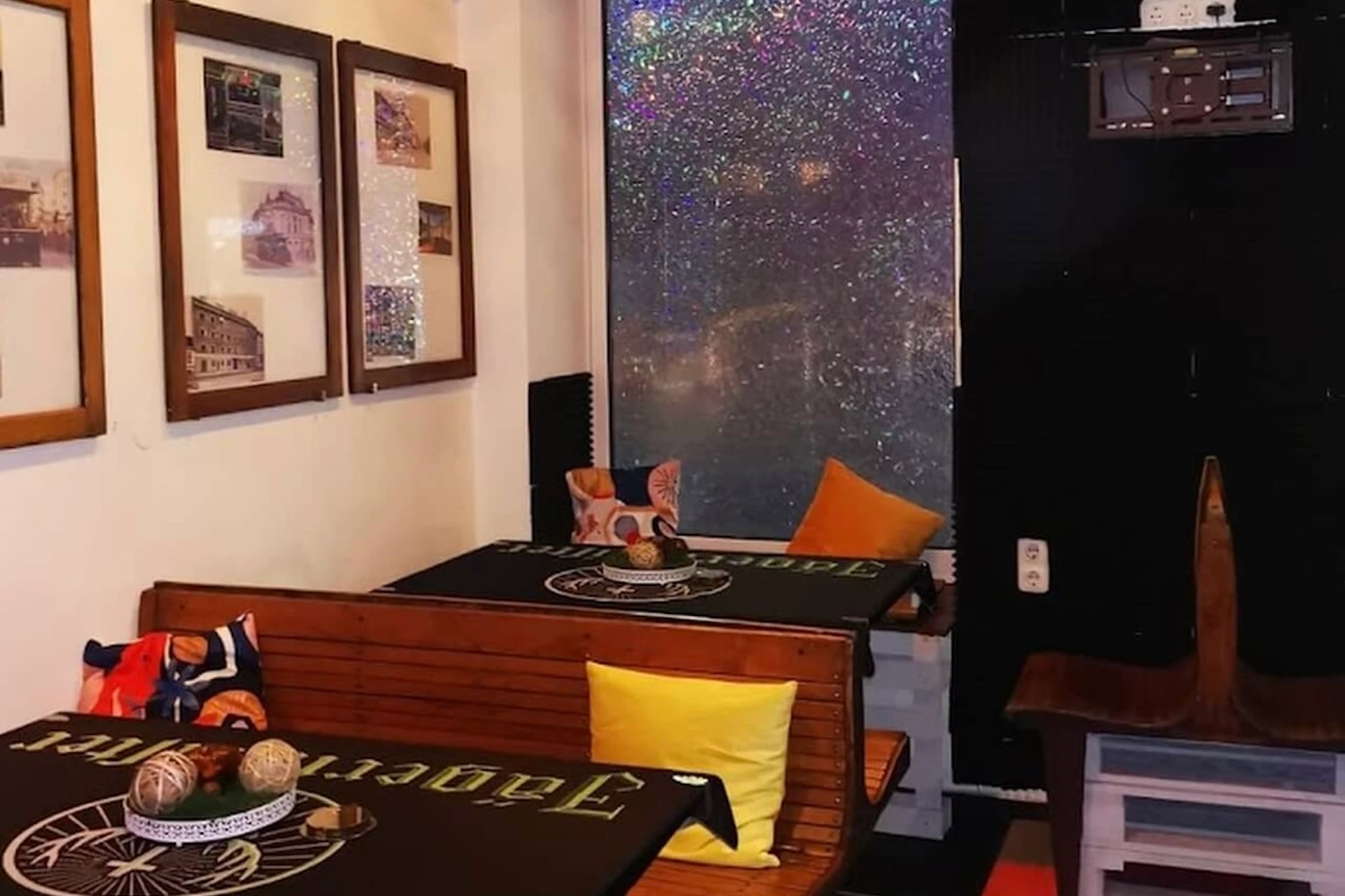 Ein gemütliches Café-Interieur mit Holzbänken, schwarzen Tischplatten, bunten Kissen, gerahmten Fotos an der Wand und einem funkelnden Fenster im Hintergrund. Auf den Tischen stehen dekorative Gegenstände.