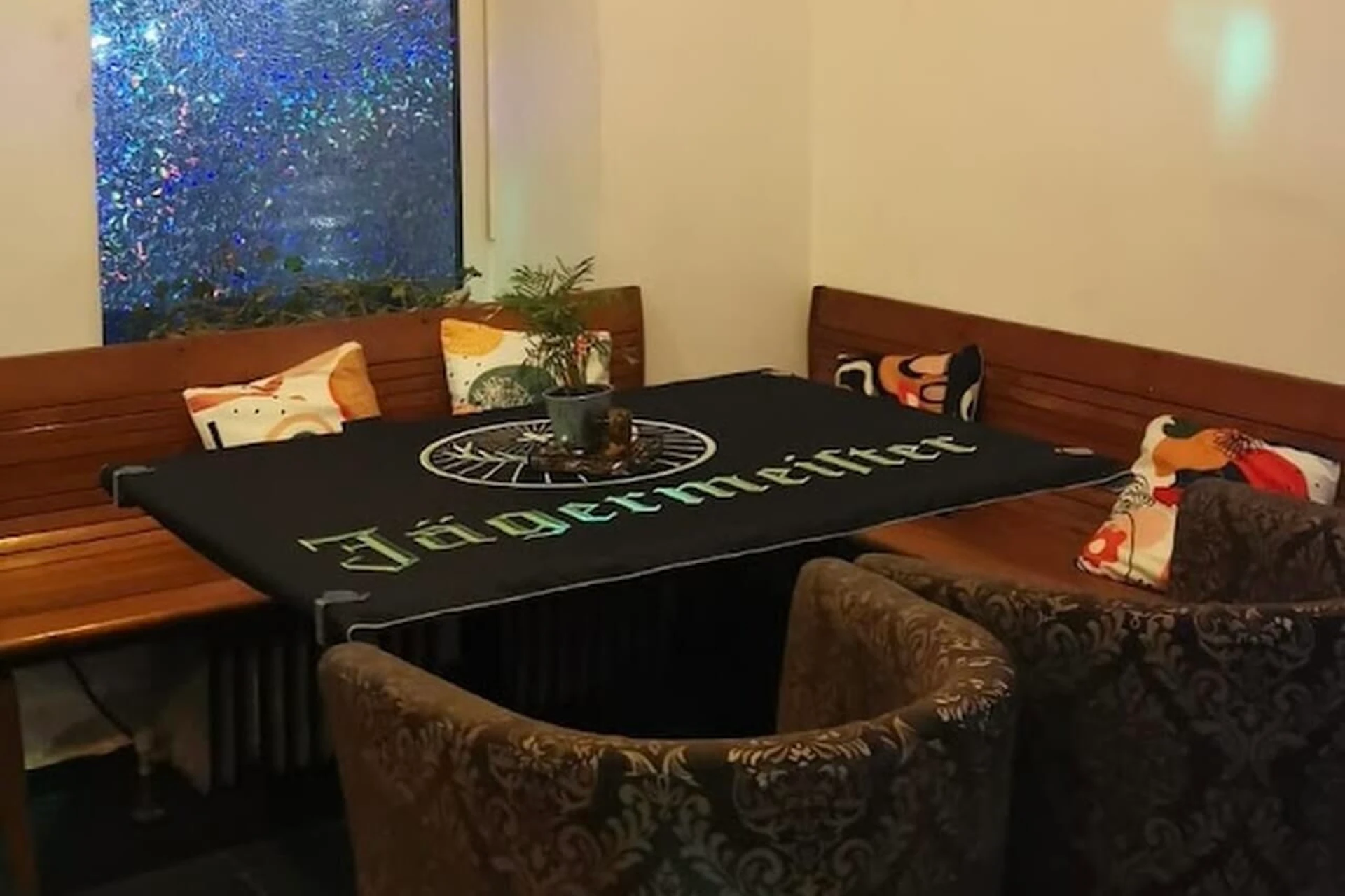 Eine gemütliche Sitzecke mit Holzbänken und gemusterten Stühlen um einen Tisch, der mit einem schwarzen Jägermeister-Tuch bedeckt ist, dekoriert mit Topfpflanzen und bunten Kissen in der Nähe eines Fensters.