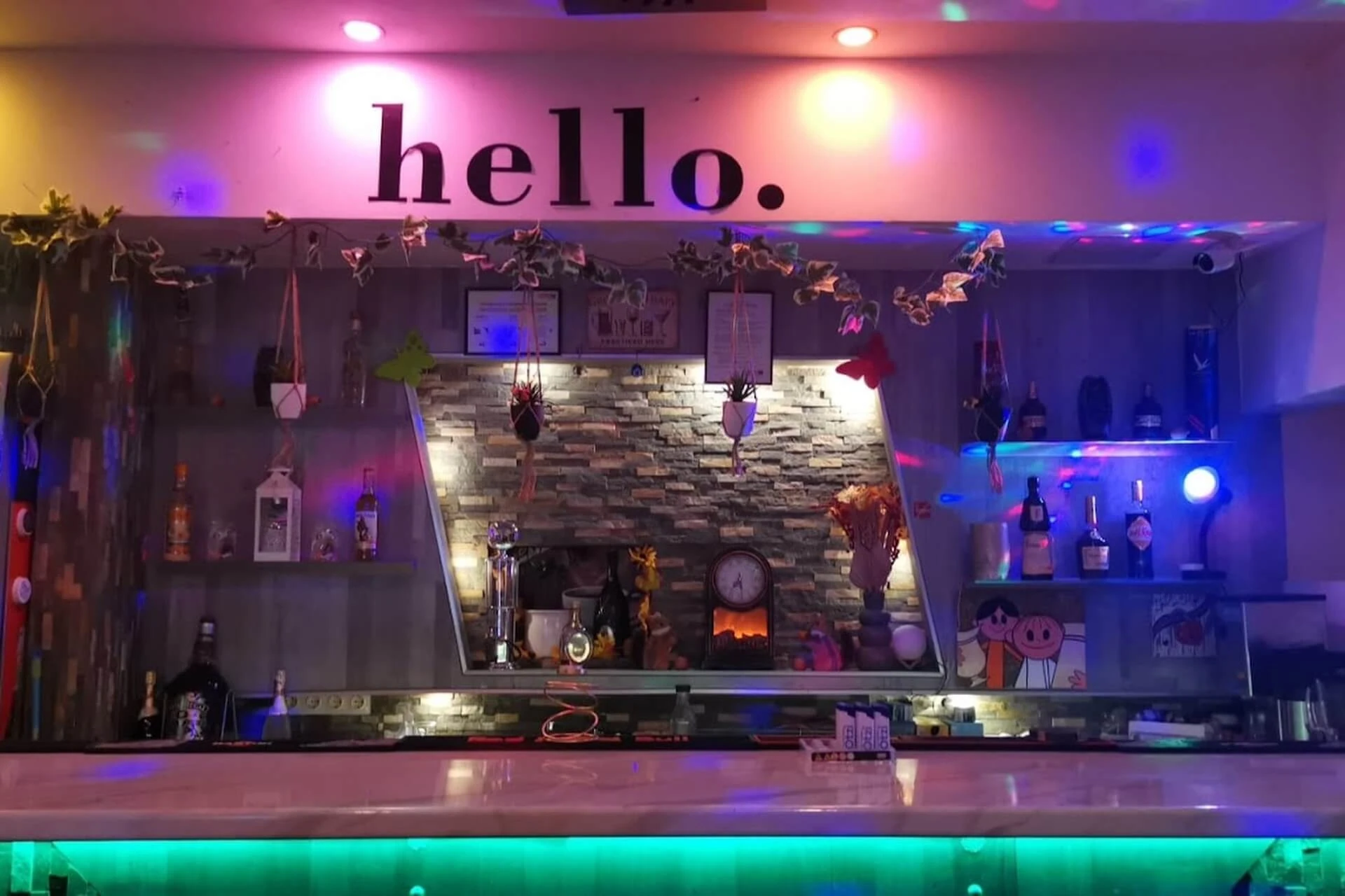 Eine bunte Bar mit Flaschen, hängenden Pflanzen und festlichen Lichtern. Das Wort "Hallo" steht in fetten schwarzen Buchstaben über der Bar, die mit verschiedenen kleinen Objekten dekoriert und mit rosa und grünen Lichtern beleuchtet ist.
