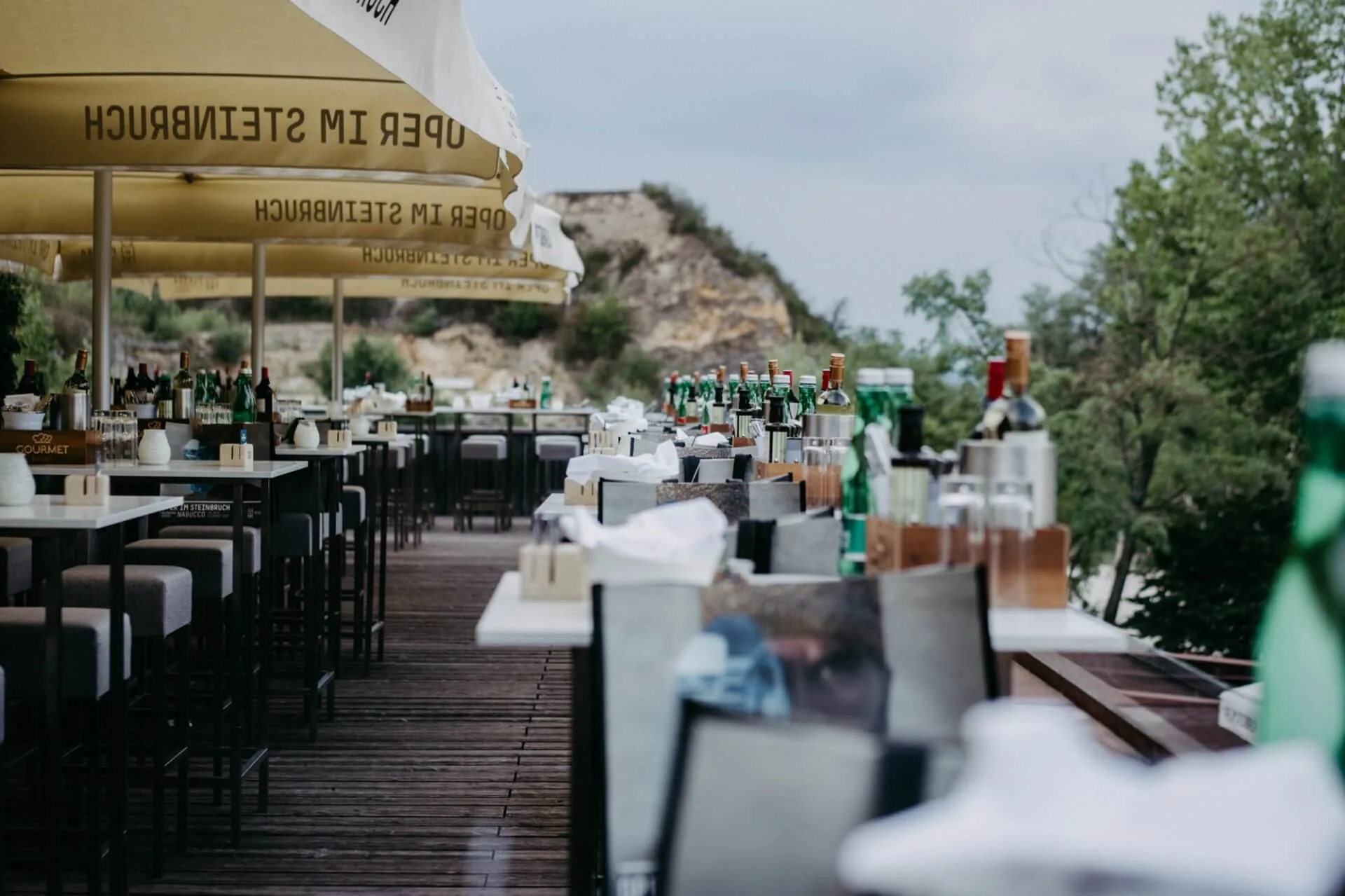 Restaurantterrasse im Freien mit Tischen zum Essen, Flaschen, Gläsern und Sonnenschirmen als Schattenspender. Der Bereich bietet einen Blick auf eine malerische, grüne Landschaft mit Bäumen und felsigen Hügeln im Hintergrund.
