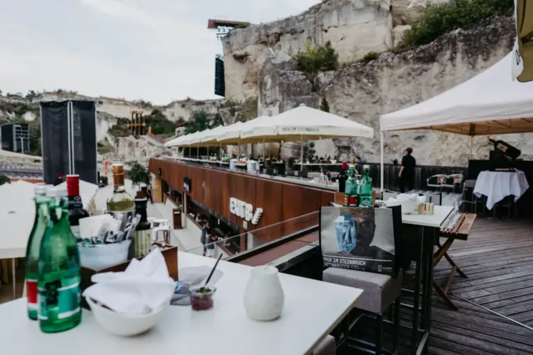 Ein Freiluftrestaurant mit Tischen zum Essen, die von großen Sonnenschirmen beschattet werden, direkt an den Felsen gelegen. Flaschen, Gläser und Speisekarten sind auf den Tischen und an der Bar zu sehen, während das Personal im Hintergrund steht.