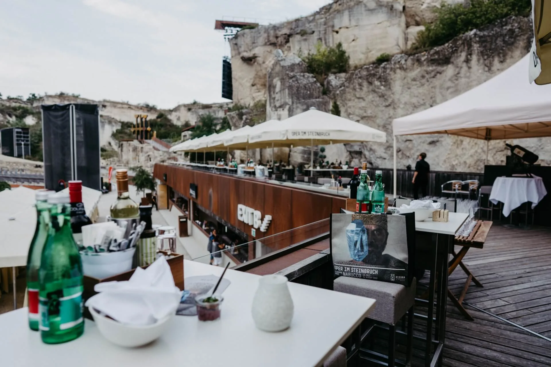 Ein Freiluftrestaurant mit Tischen zum Essen, die von großen Sonnenschirmen beschattet werden, direkt an den Felsen gelegen. Flaschen, Gläser und Speisekarten sind auf den Tischen und an der Bar zu sehen, während das Personal im Hintergrund steht.