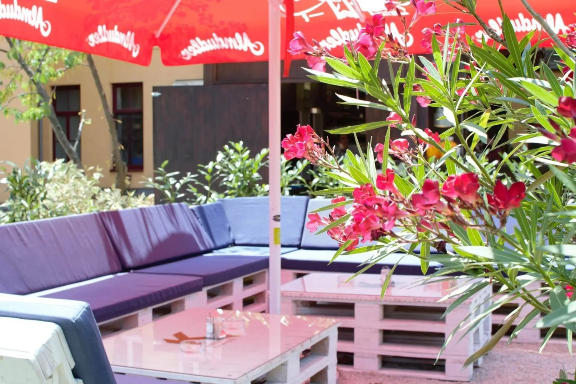 Sitzbereich im Freien mit blau gepolsterten Bänken aus Holzpaletten, einem weißen Couchtisch aus Paletten und einem großen roten Sonnenschirm. Im Vordergrund stehen rosa blühende Pflanzen, im Hintergrund Grünzeug.