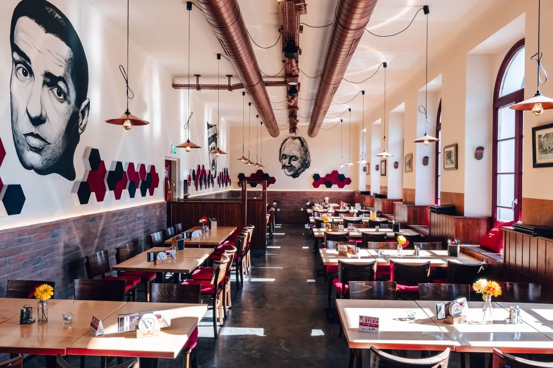 Ein geräumiges Restaurant mit Holztischen und roten Stühlen, dekoriert mit großen schwarz-weißen Wandbildern von Gesichtern an den Wänden, Hängelampen und sechseckiger Wandkunst. Das Sonnenlicht strömt durch die hohen Fenster.