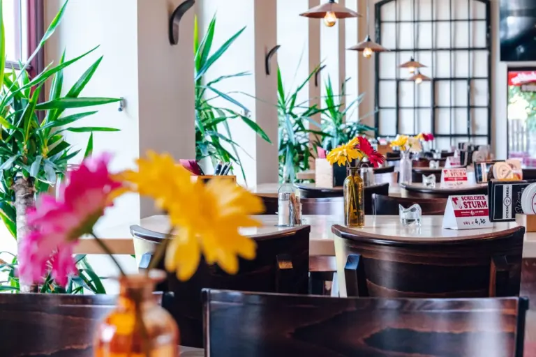 Ein helles, modernes Restaurant mit Holztischen und -stühlen, Vasen mit bunten Blumen und Grünpflanzen an den Fenstern. Natürliches Licht durchflutet den Raum und schafft eine einladende und frische Atmosphäre.