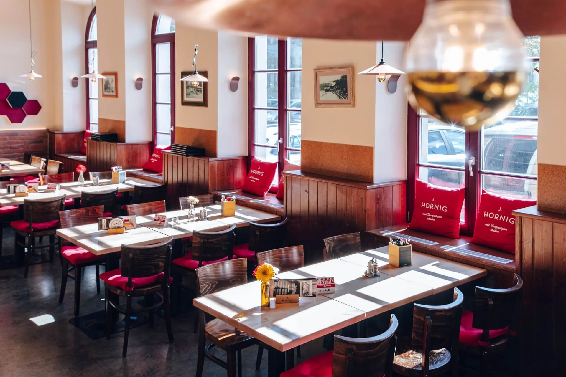 Ein helles, gemütliches Restaurant mit Holztischen und -stühlen, roten Kissen, Sonnenblumen in Vasen, großen Fenstern, die das Sonnenlicht hereinlassen, und von der Decke hängenden Lampen.