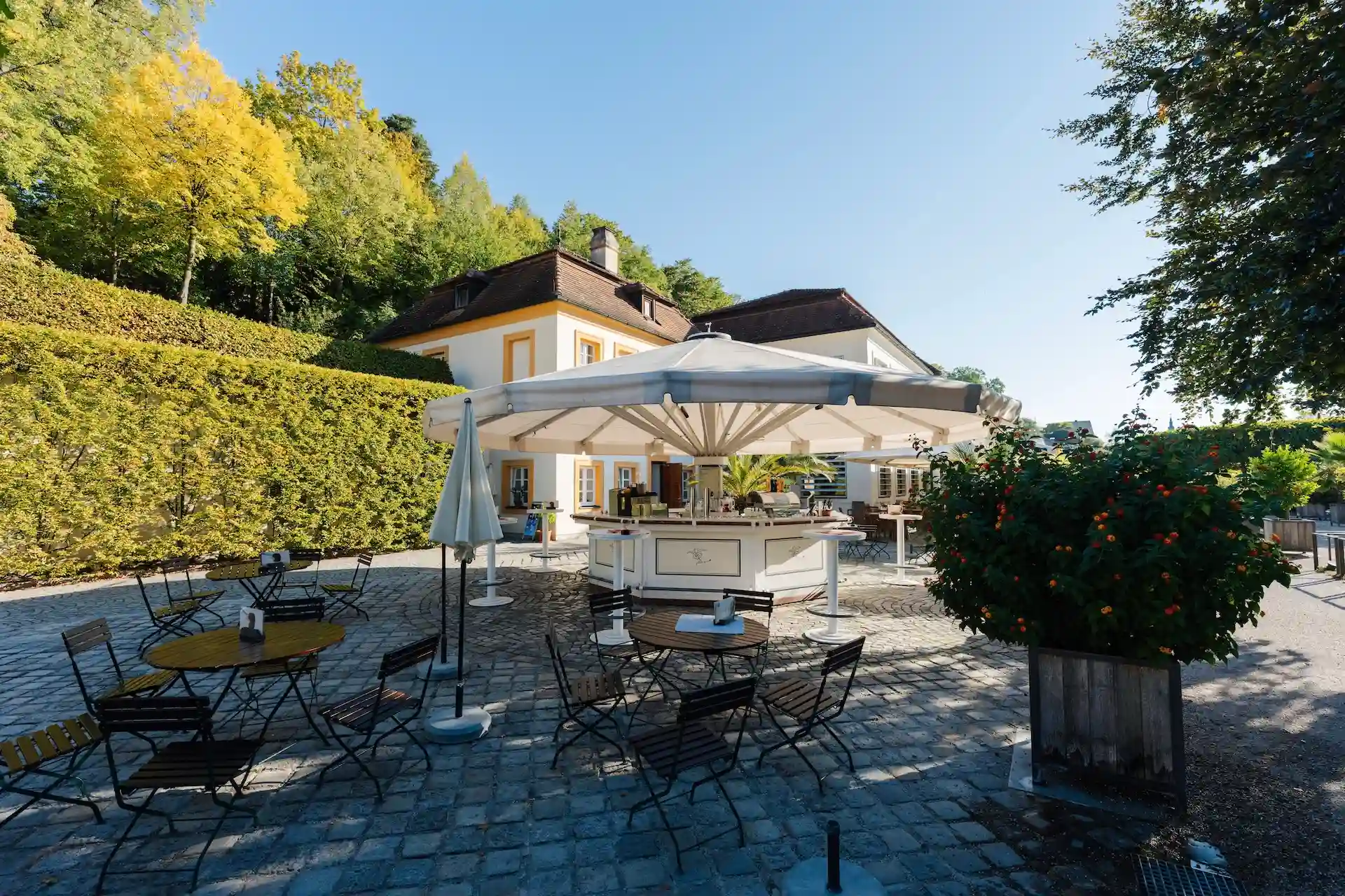 Terrasse eines Restaurants im Freien mit Tischen und Stühlen unter einem großen weißen Sonnenschirm vor einem Gebäude, das sich an einen Hang schmiegt.