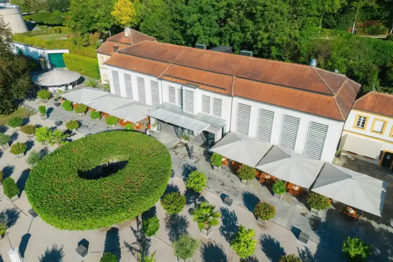 Stiftsrestaurant Melk Luftaufnahme eines langgestreckten Gebäudes mit rotem Ziegeldach und einer Terrasse mit Sitzgelegenheiten im Freien, die von Sonnenschirmen beschattet wird und an eine große, kreisförmige Hecke grenzt.