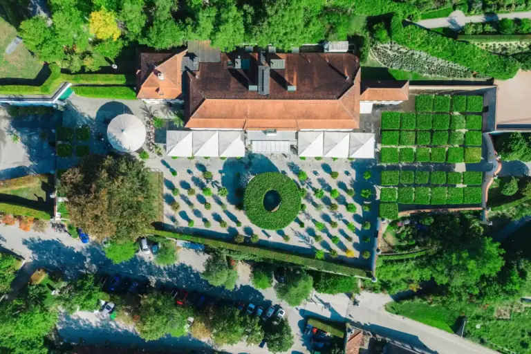 Eine Luftaufnahme eines großen Gebäudes mit Terrakottadach, das über einen gepflegten Innenhof mit Baumreihen, einen kreisförmigen Garten und einen nahegelegenen Parkplatz verfügt.